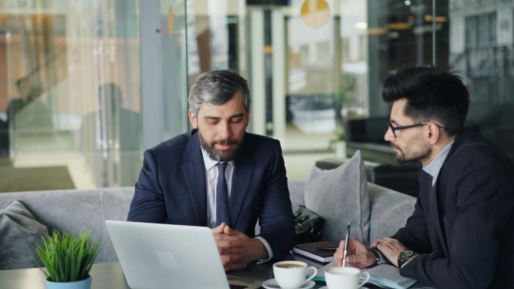 pexels-photo-23496863-23496863 Two businessmen in suits discussing work at a cafe with laptops and coffee.